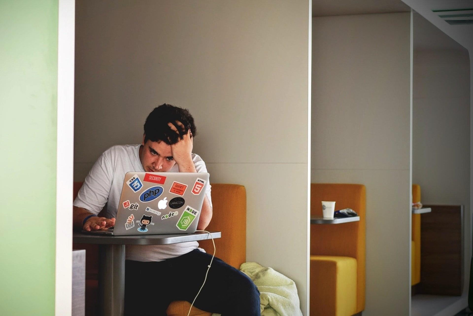 Man sitting at a table working on his resume on his laptop and holding his forehead in frustration