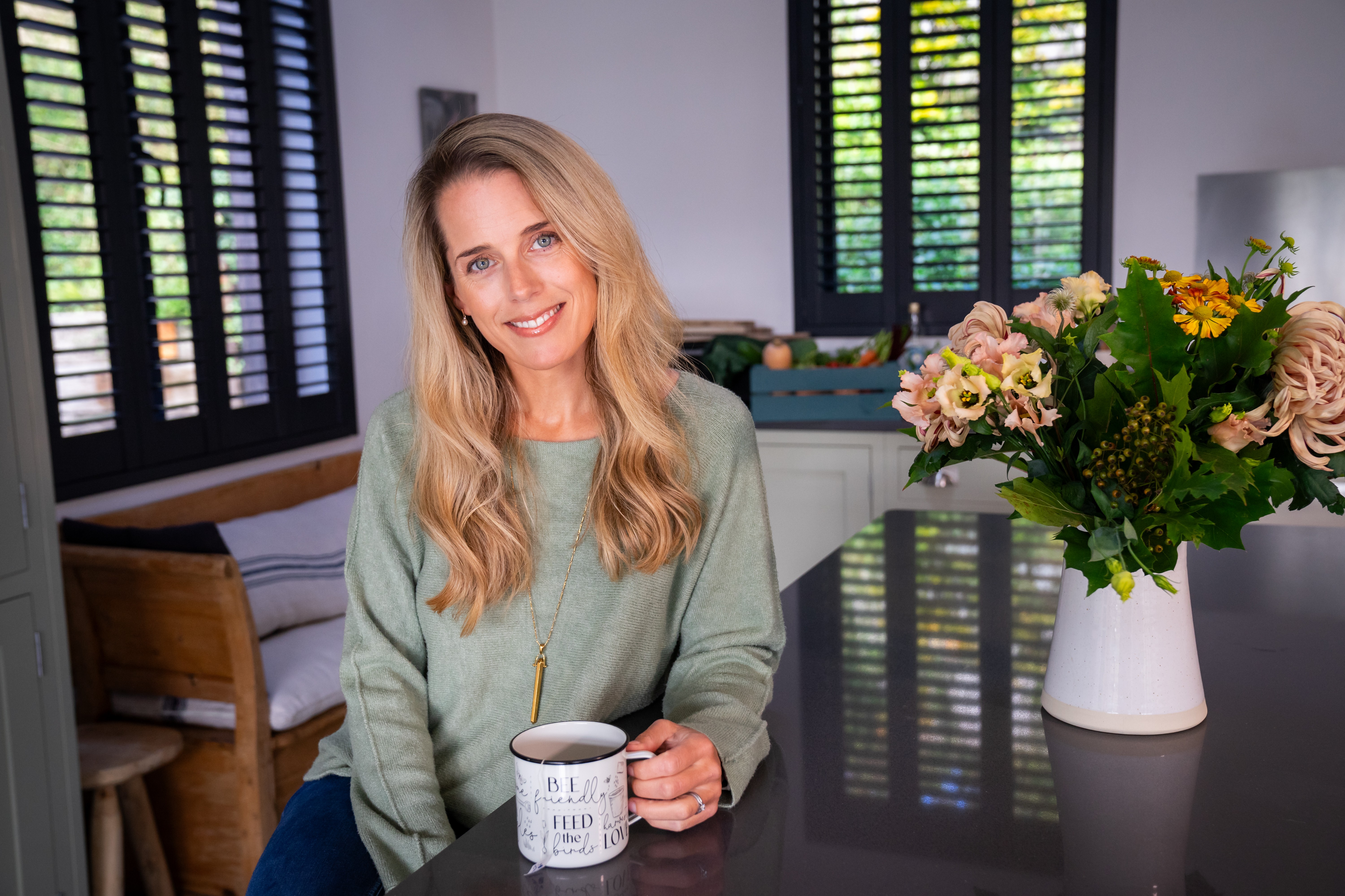 Ellie White, women’s wellness coach, sitting in her kitchen holding a coffee mug beside fresh flowers.