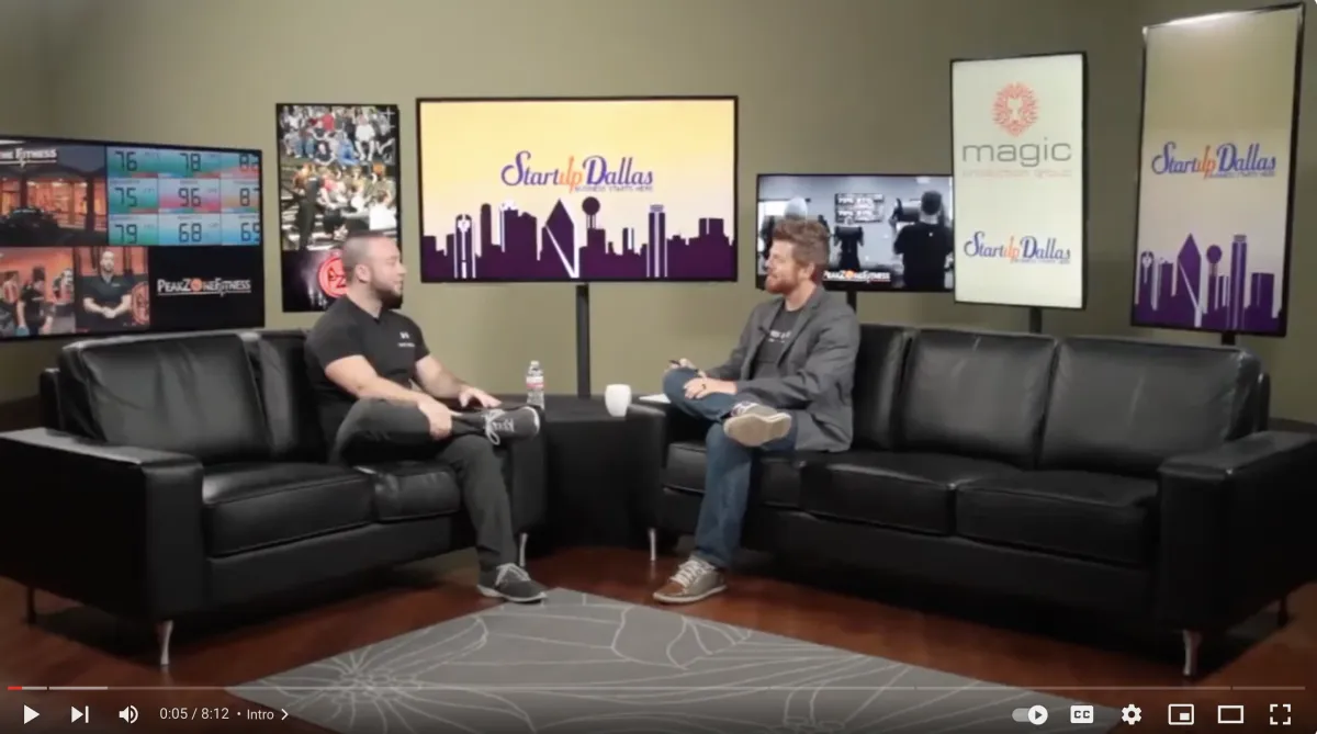 Two men seated on black couches discussing fitness, with Peak Zone Fitness branding visible on screens in the background, highlighting Coach Donny Day's media presence and fitness expertise.