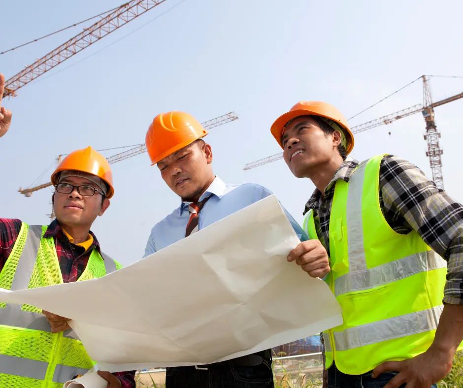 Three construction workers in hard hats and safety vests review blueprints on a job site with cranes in the background.
