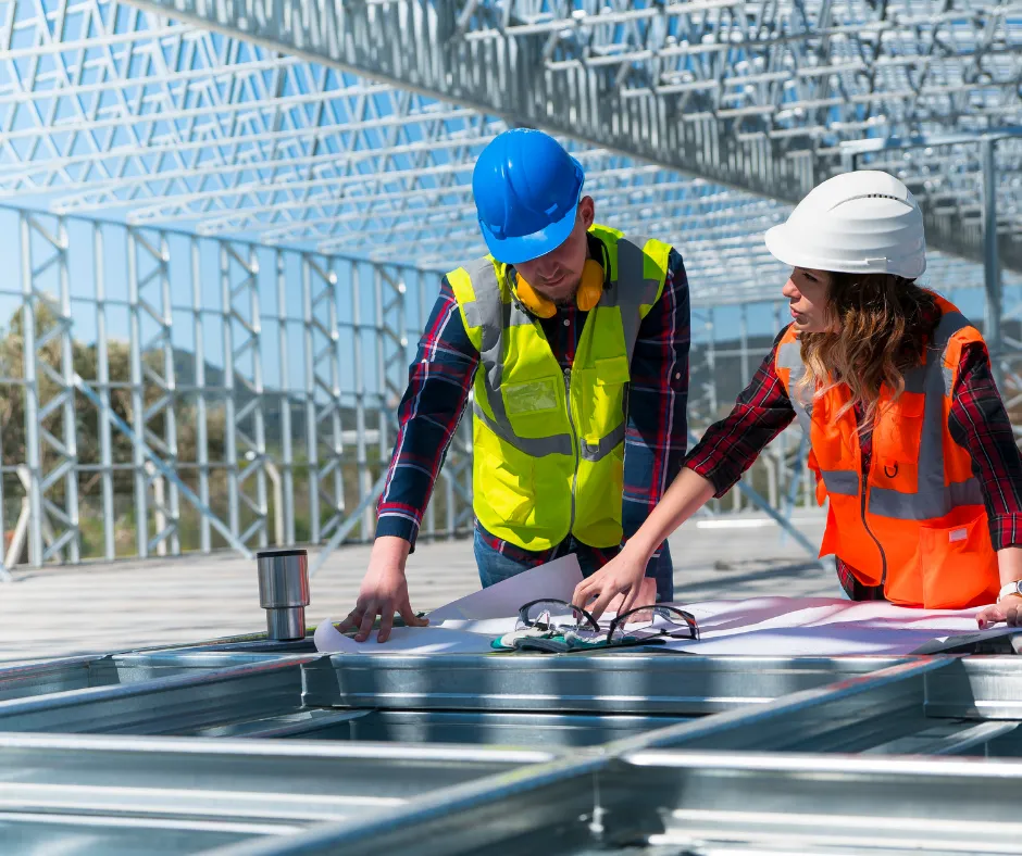 Two construction workers in safety vests examine blueprints on metal framework under a clear sky.