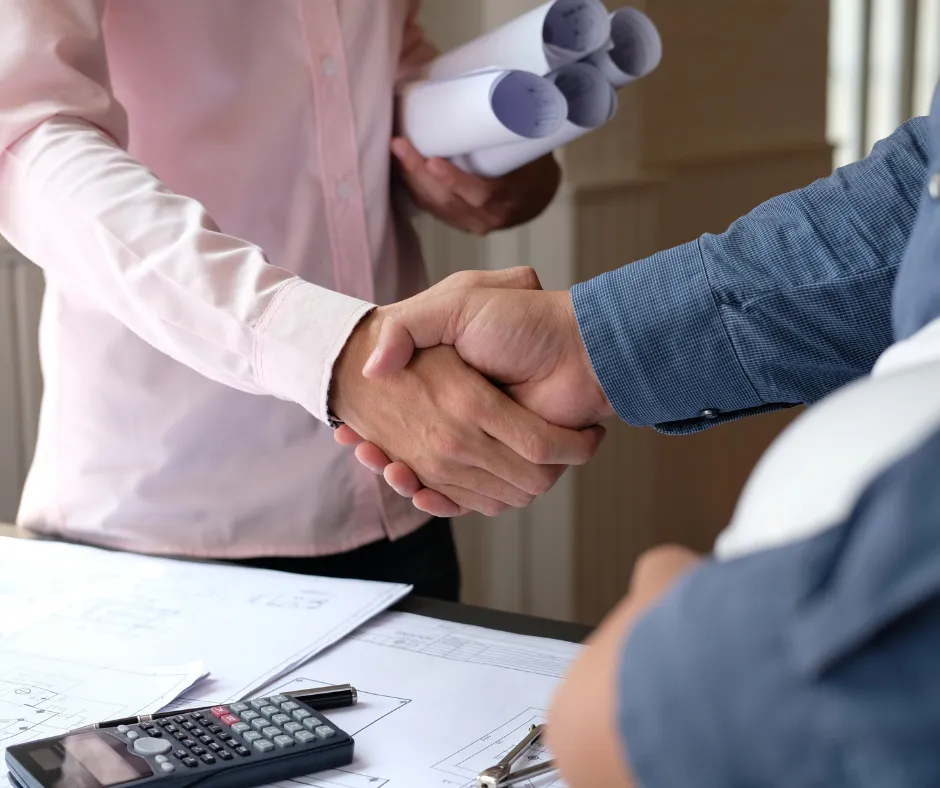 Two professionals shake hands over blueprints and a calculator on a desk