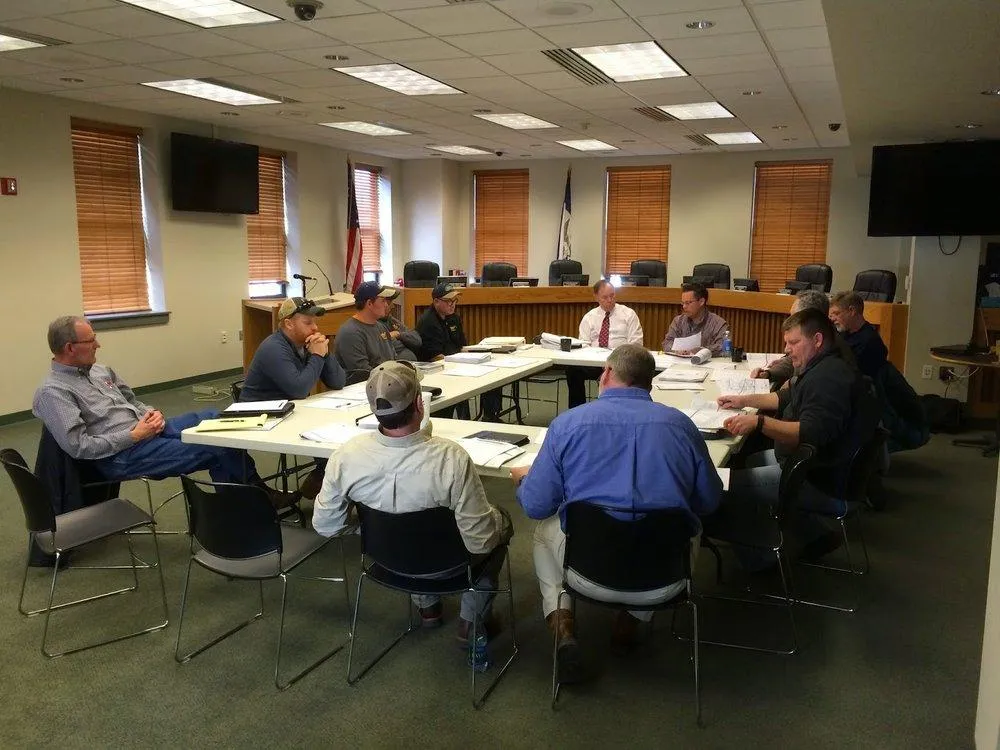 A group of men sits around a table in a conference room, discussing plans with papers and documents.
