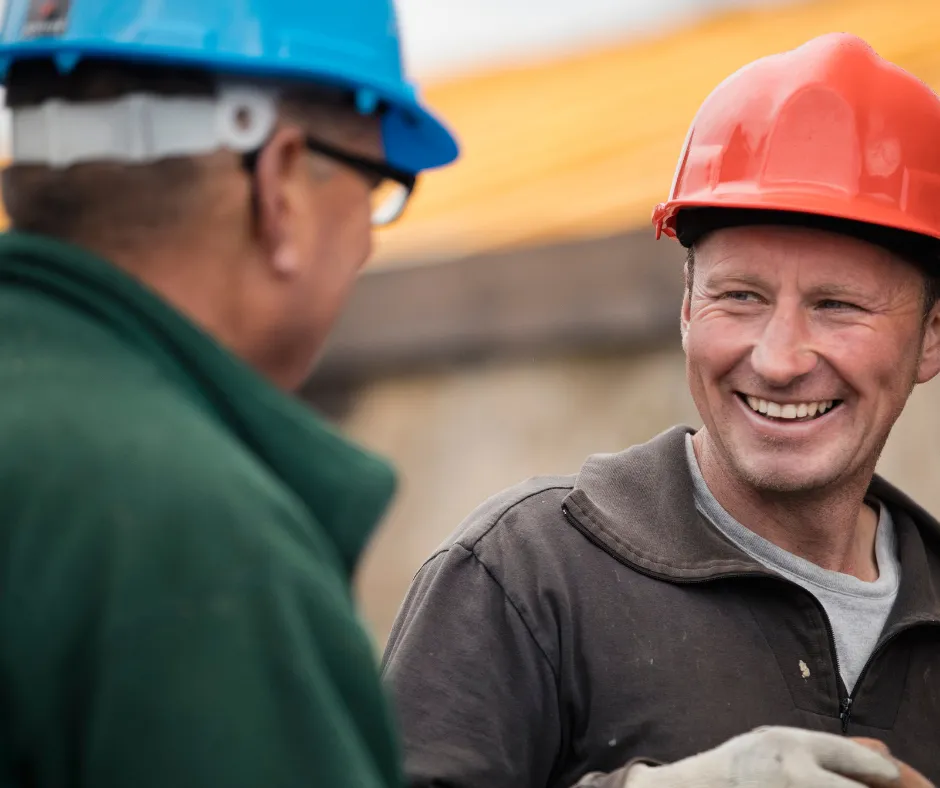 Two construction workers engage in conversation, one wearing a blue hard hat and the other in a red hard hat.