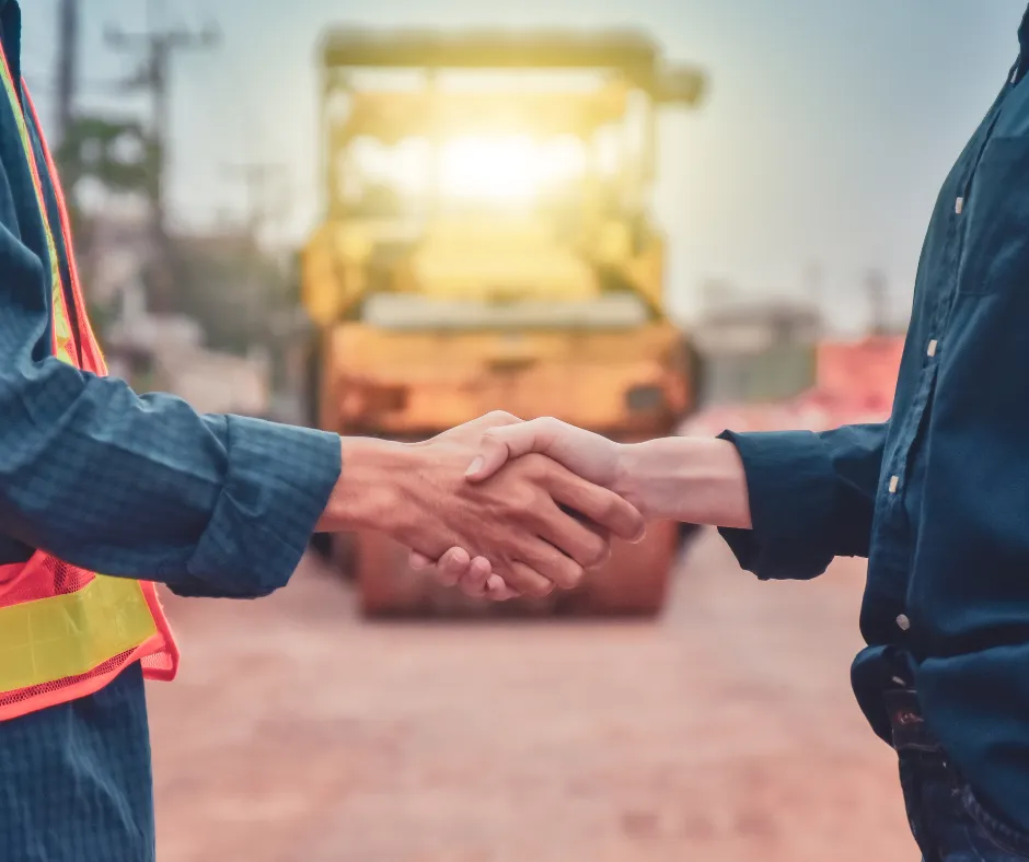 Two individuals shake hands in a construction setting, with a yellow steamroller visible in the background.