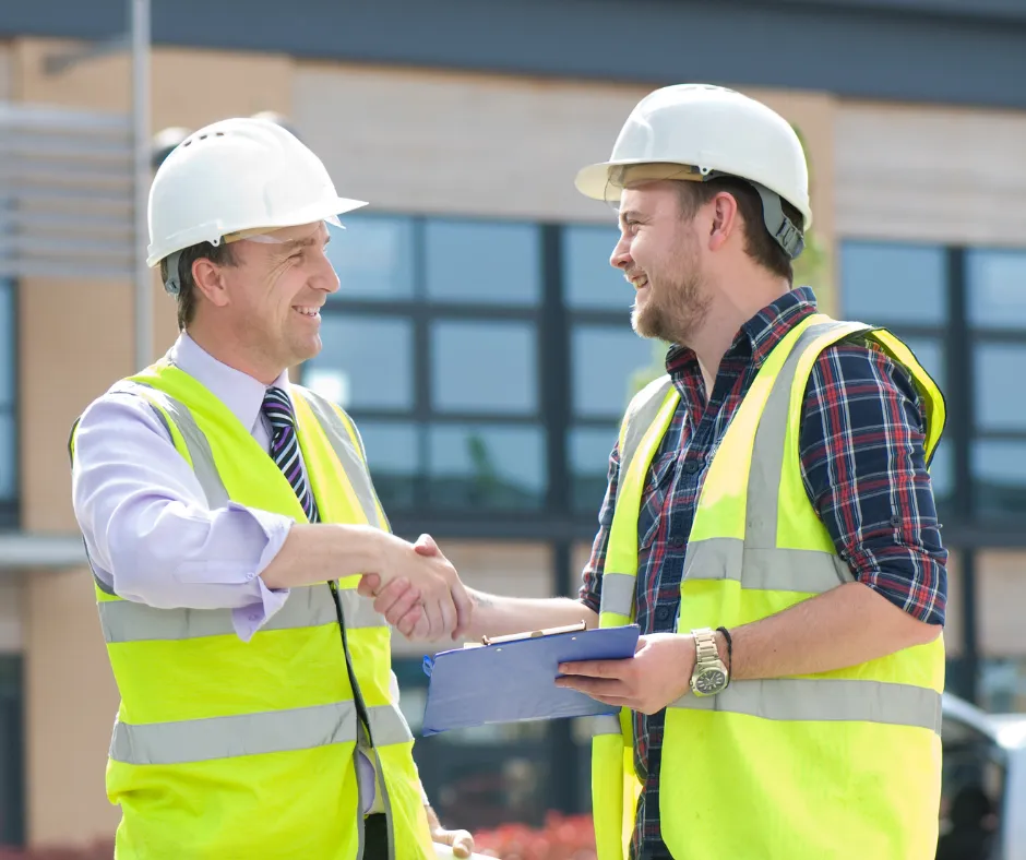 Two construction workers in safety vests and helmets shake hands in front of a modern building