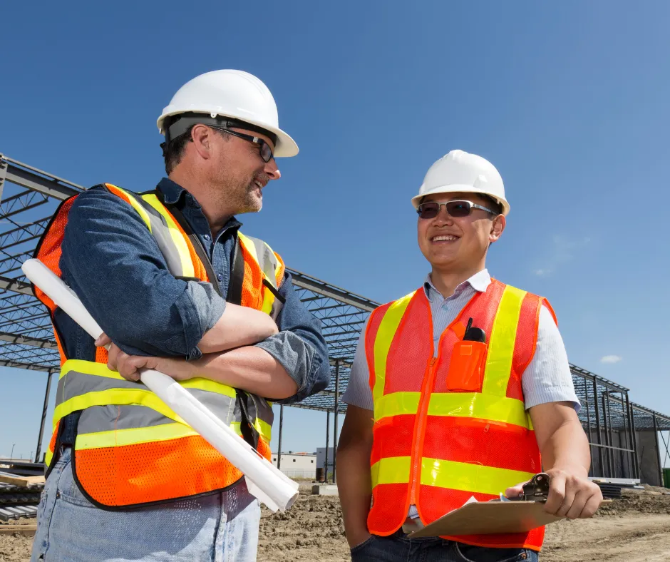 Two construction workers in safety vests and hard hats stand together on a building site, discussing plans with a steel structure