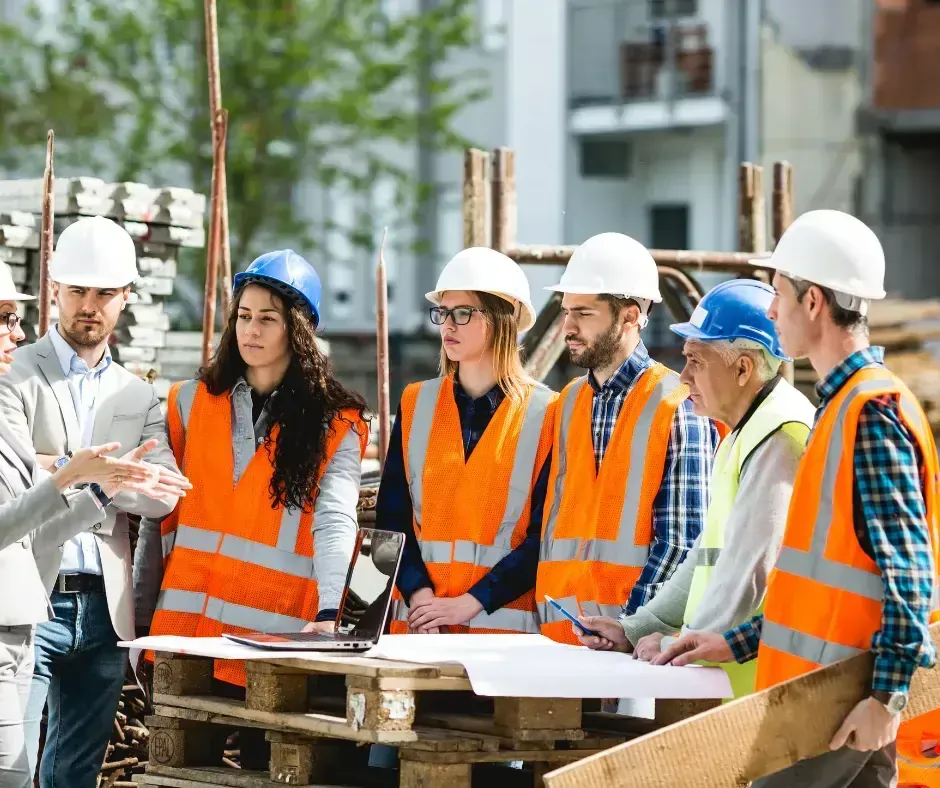 A diverse group of construction workers in safety vests and hard hats discusses plans at a job site