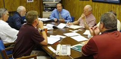 A group of men sits around a table in a conference room, discussing plans with papers and documents.