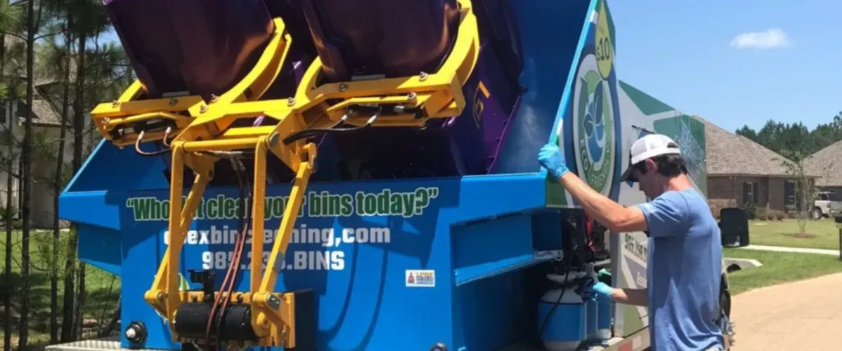 Technician operating trash bin cleaning truck while bins are lifted for washing