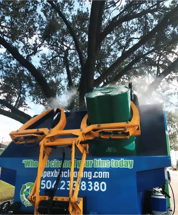 A green bin is being cleaned with steam by a blue and yellow bin cleaning truck under a large tree.