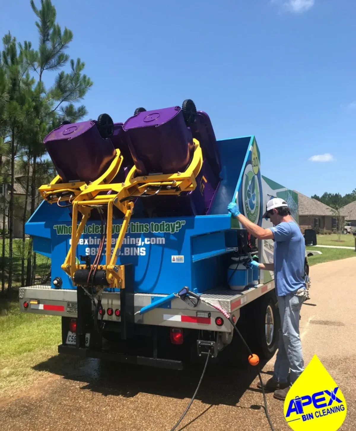 A man in a hat and blue gloves operates a blue truck cleaning two purple garbage bins on a sunny day.
