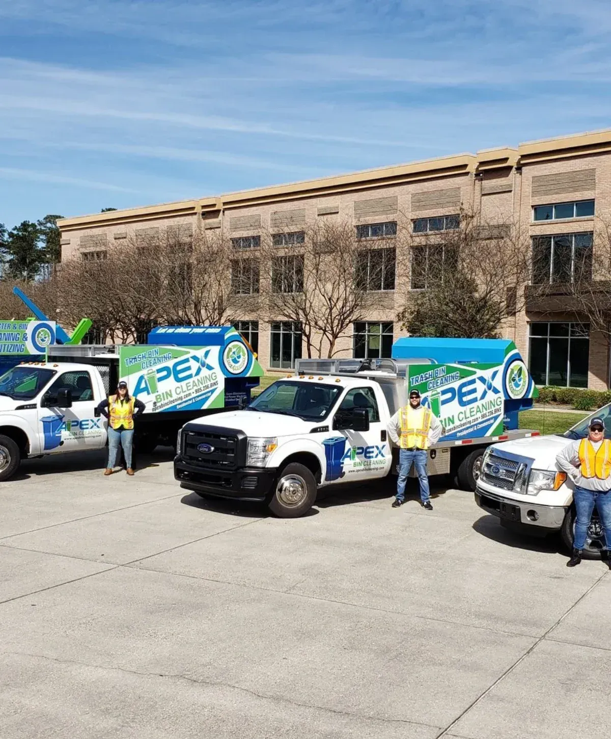 Three white Apex Bin Cleaning trucks with their crew in high-vis vests parked in front of a brick building.