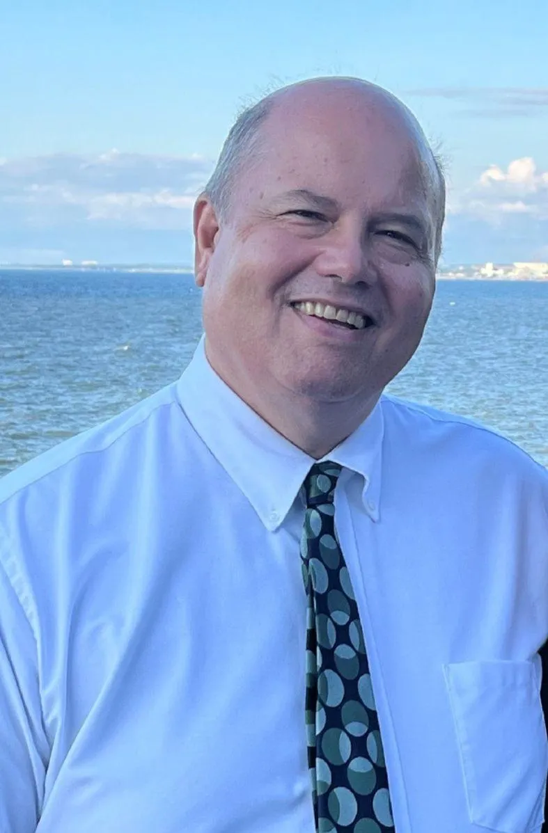 Portrait of a smiling male financial planner in his 50s, wearing a navy suit and glasses, standing in front of a bookshelf filled with financial books.