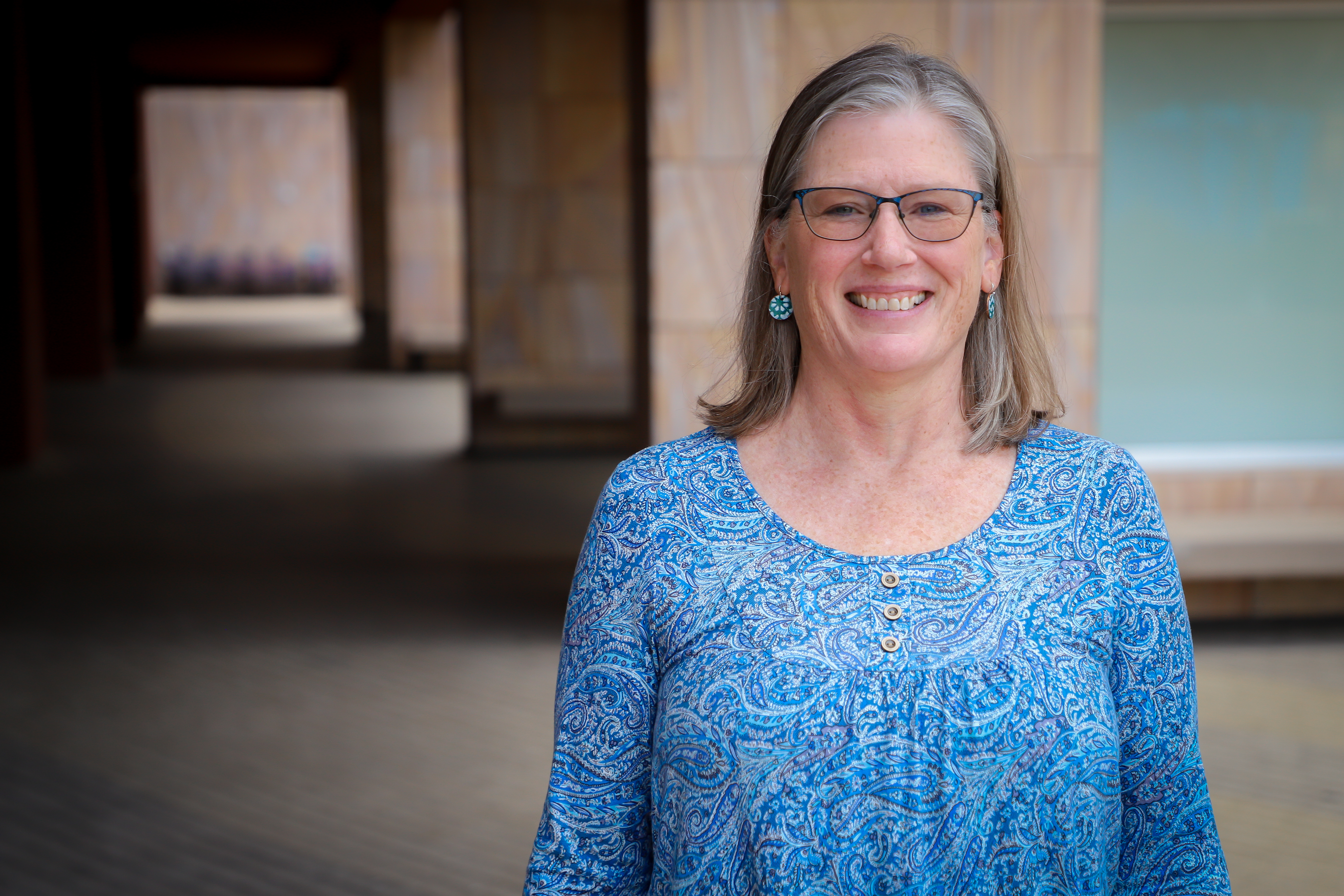 Portrait of a cheerful female Medicare specialist in her 40s, with curly hair and a teal blouse, seated at a desk with a notepad and pen.