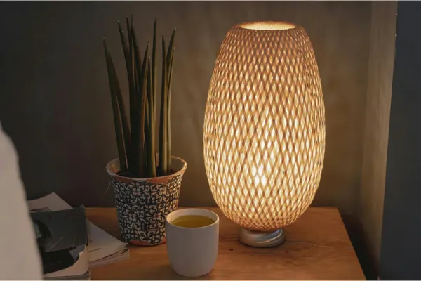 A woven lamp glowing warm in a dim bedroom, with tea and a plant on the nightstand, evening wind-down lighting.