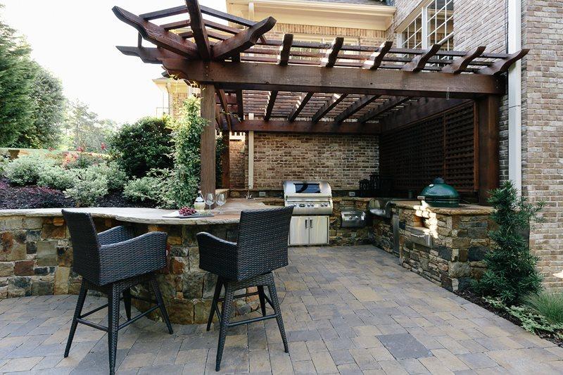 Outdoor kitchen and patio with stone counters, bar stools, grill, and a wooden pergola attached to a brick house.