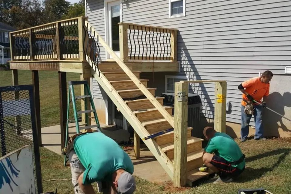 Workers constructing a raised wooden deck with a staircase leading down to the yard, attached to a house.