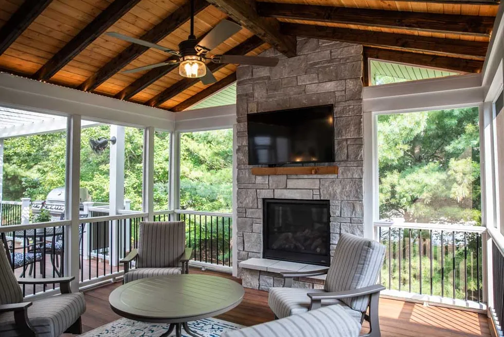 Covered deck with wooden ceiling, ceiling fan, stone fireplace, mounted TV, and outdoor seating area with chairs and table