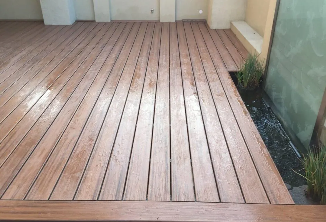 Modern wooden deck with evenly spaced planks, next to a small water feature and plants along the edge.