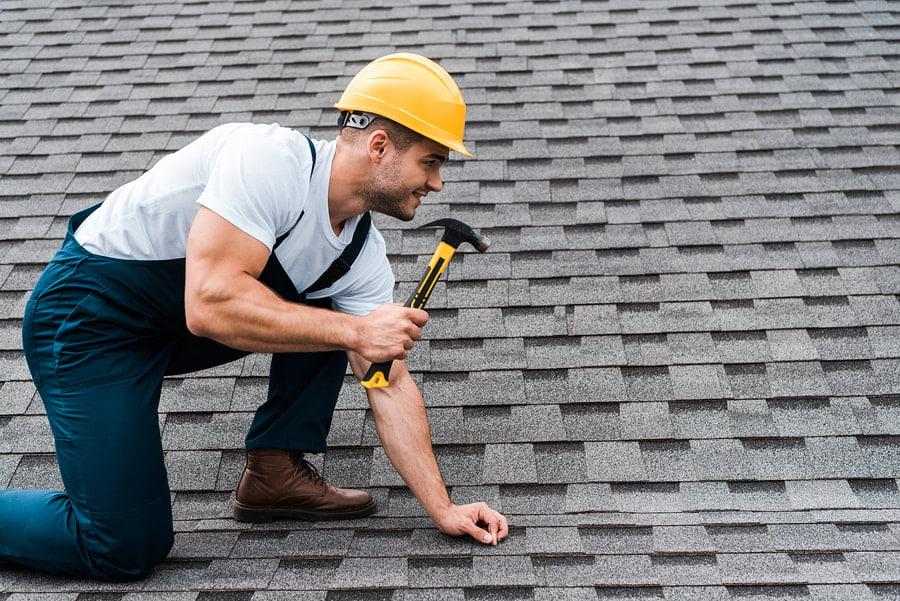 Man in a yellow hard hat and blue overalls kneeling on a shingled roof, using a hammer. He appears focused and engaged in roofing work.