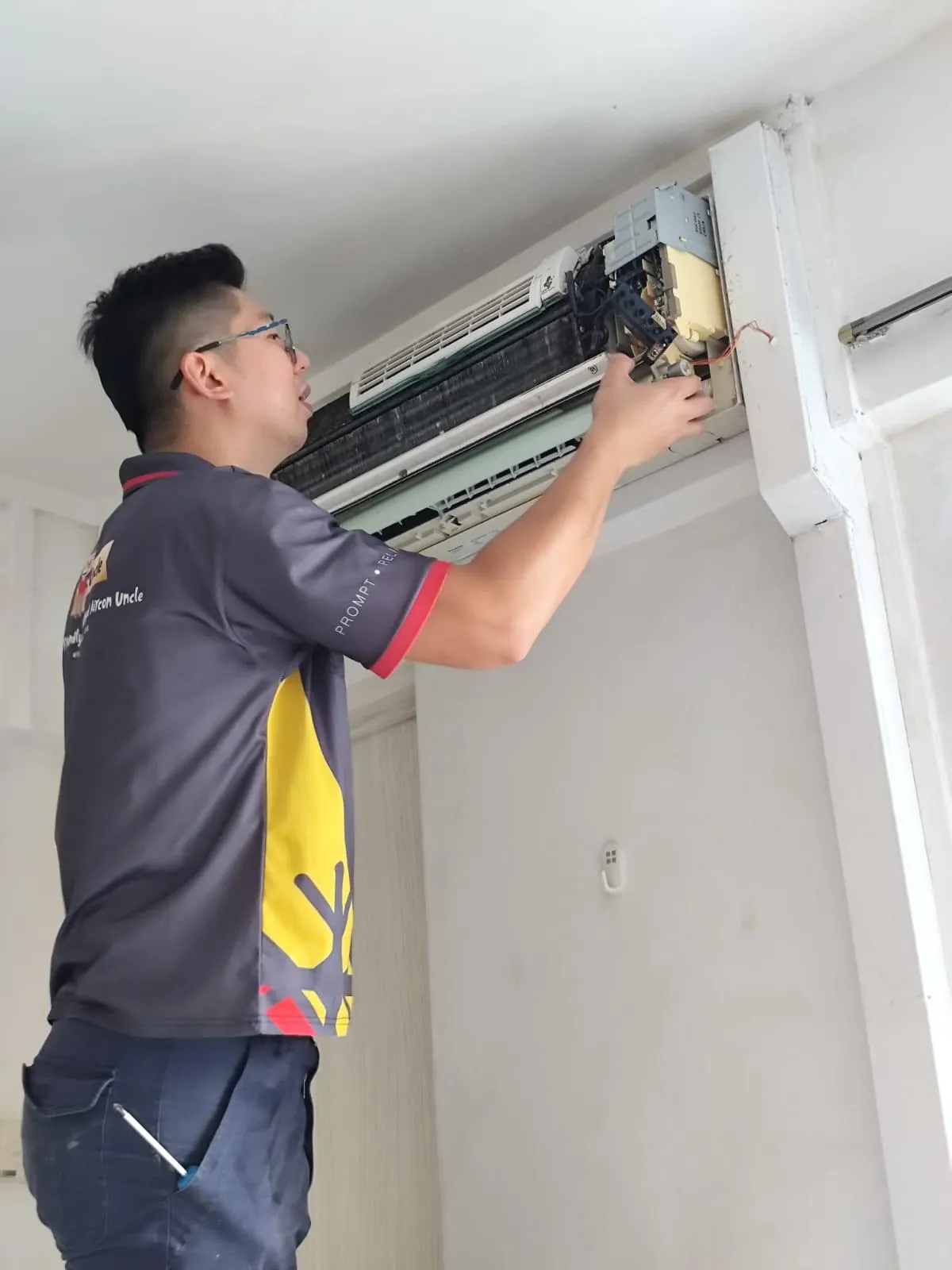 Technician performing aircon servicing on a wall-mounted unit checking drain pan, standing on a ladder to clean the interior components for optimal airflow