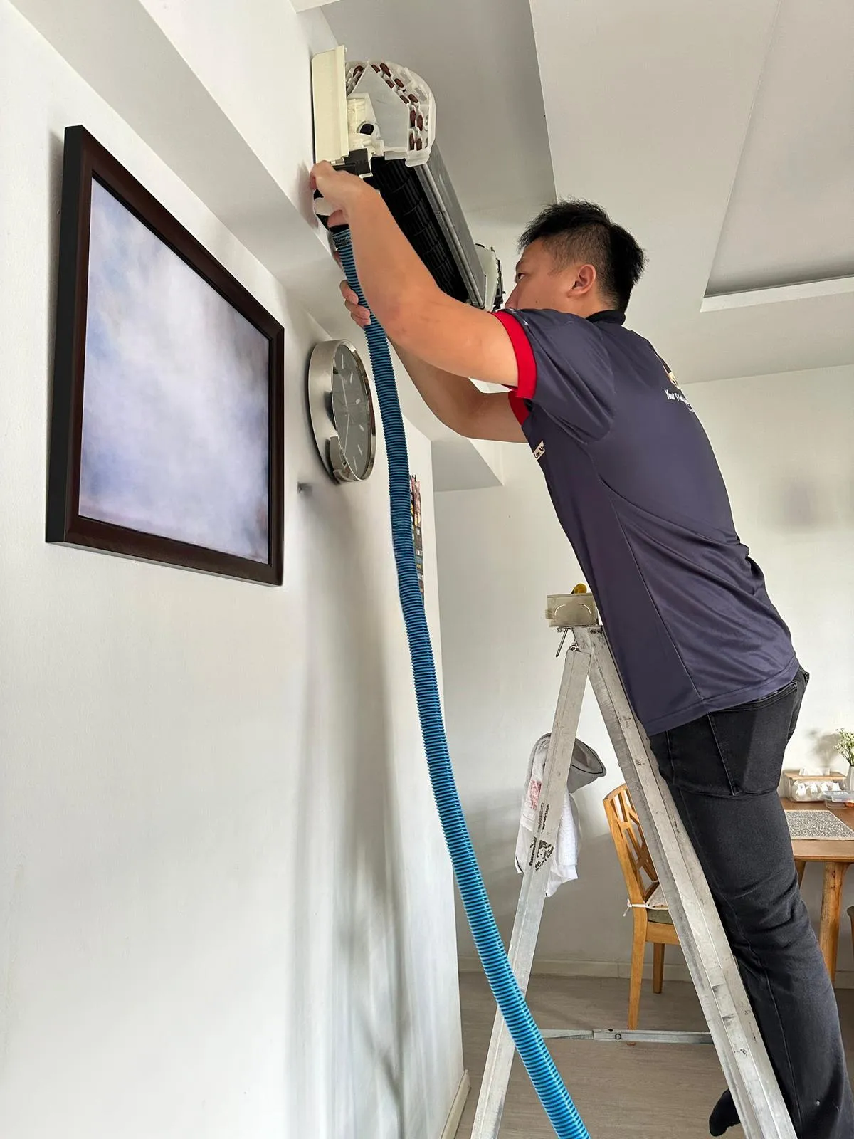 Technician performing aircon servicing on a wall-mounted unit using a vacuum hose, standing on a ladder to clean the interior components for optimal airflow.