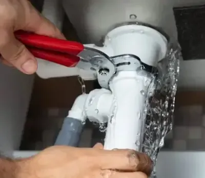 Burst pipe spraying water under sink while plumber tightens fitting with red pliers