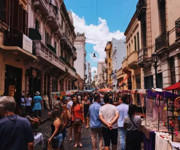 Bustling street market in San Telmo, Buenos Aires, with people shopping at colorful vendor stalls along historic European-style buildings under a bright blue sky.