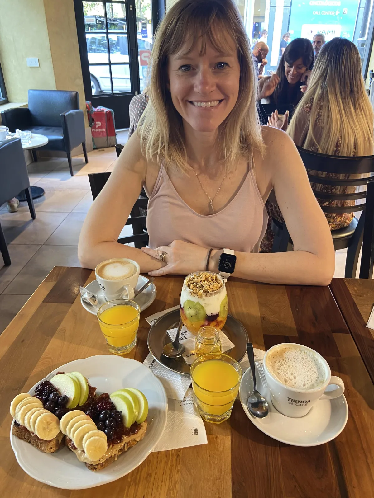 Smiling woman enjoying a healthy breakfast at a café, featuring fruit-topped toast, parfait, cappuccino, and fresh orange juice