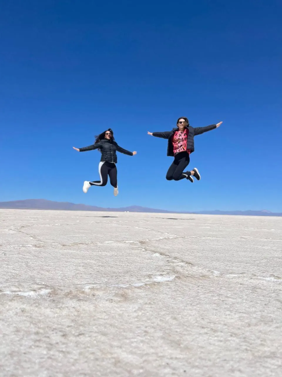 Two women jumping joyfully at Salinas Grandes salt flats in Argentina under a clear blue sky