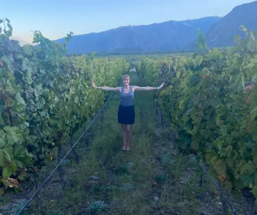 Woman standing with outstretched arms between vineyard rows at Piattelli Vineyards in Cafayate, Argentina, with scenic mountain views in the background at sunset
