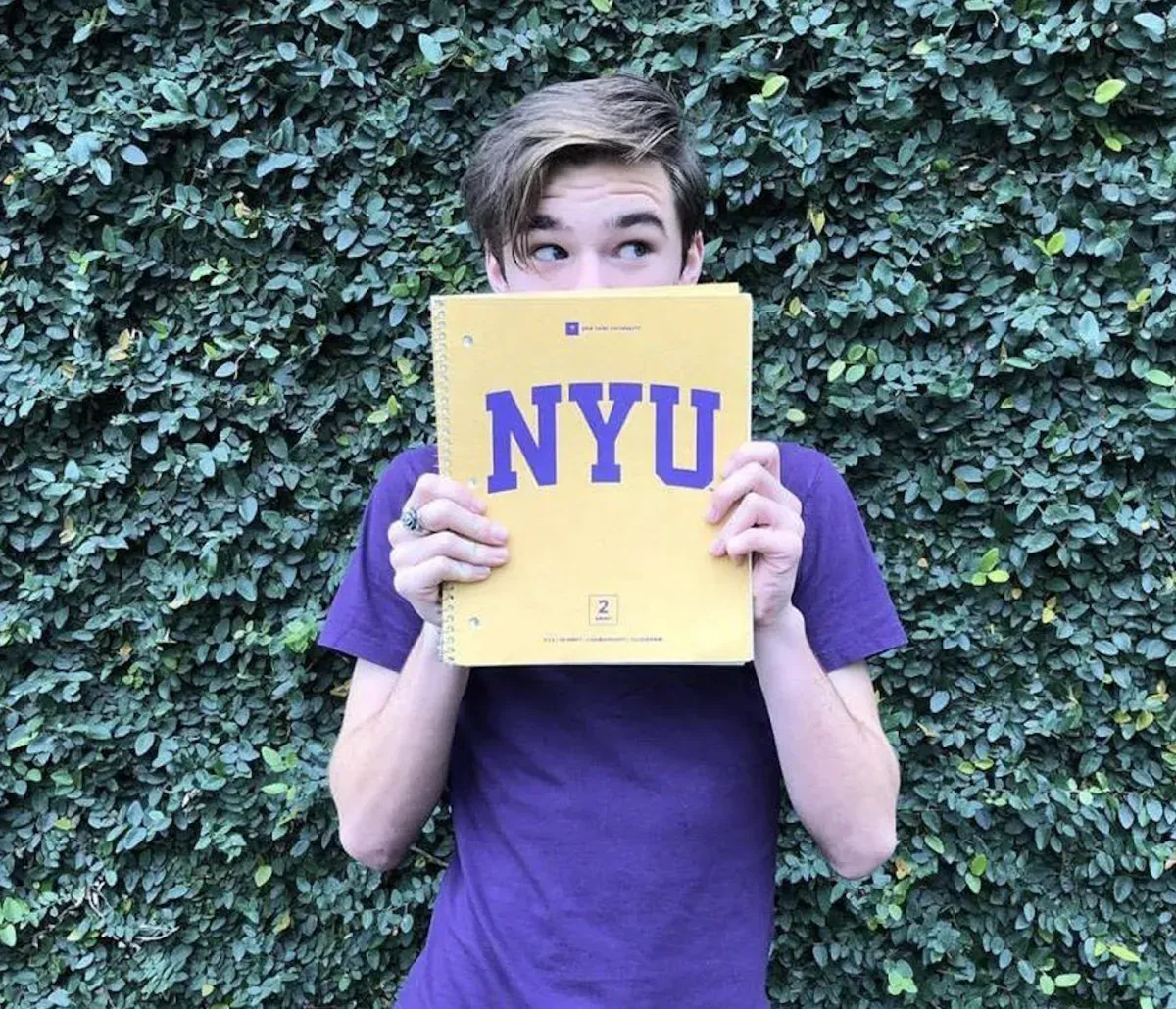 Young man holding an NYU notebook in front of his face while looking excited.