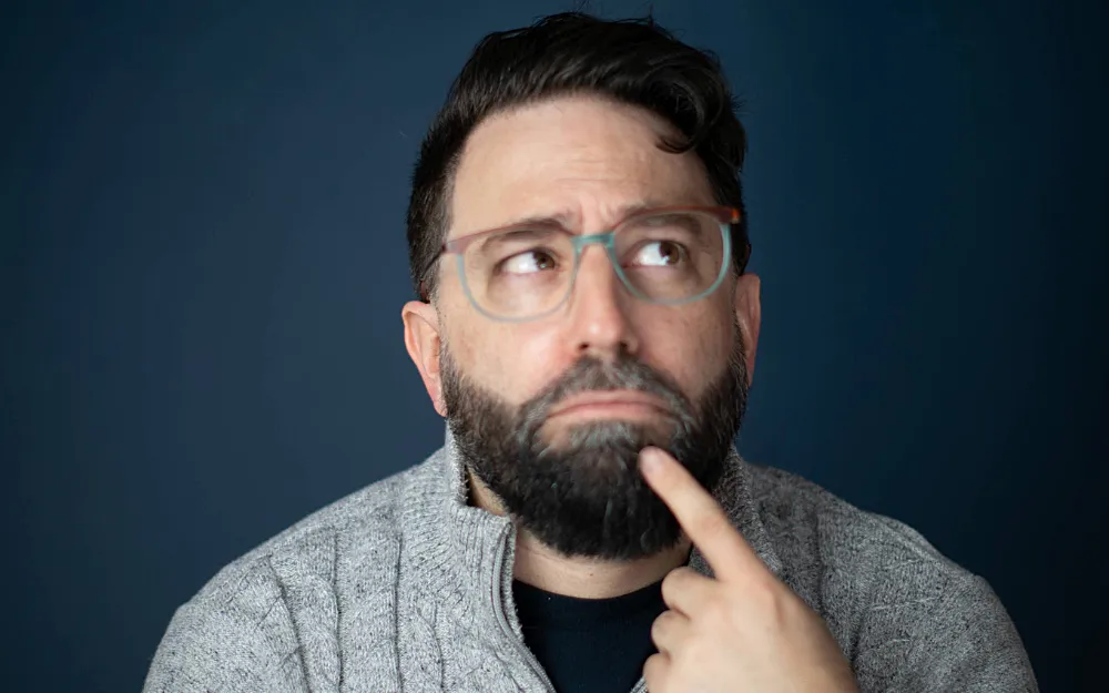 Bearded man looking inquisitive with a finger on his chin and his eyes up to the right. 