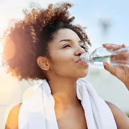 Smiling woman with curly hair and a white towel drinking water outdoors after a workout, symbolizing hydration and self-care.