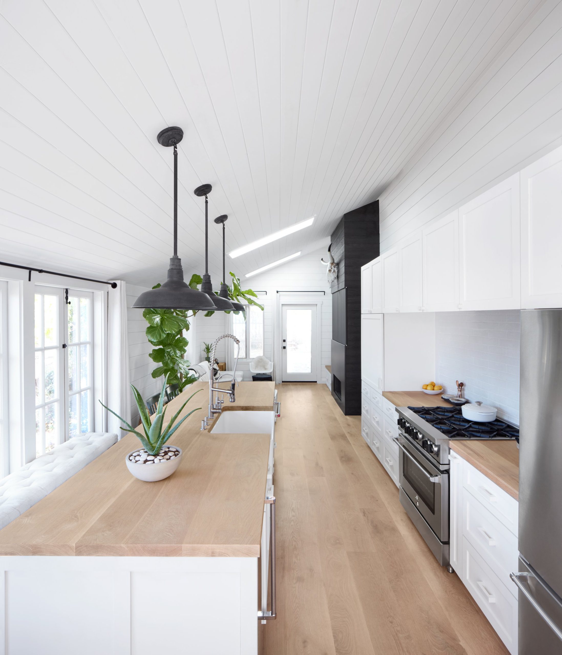 A bright, sell-ready kitchen with a long timber-topped island, white cabinetry, black pendant lights and a potted aloe, sunlight coming through French doors