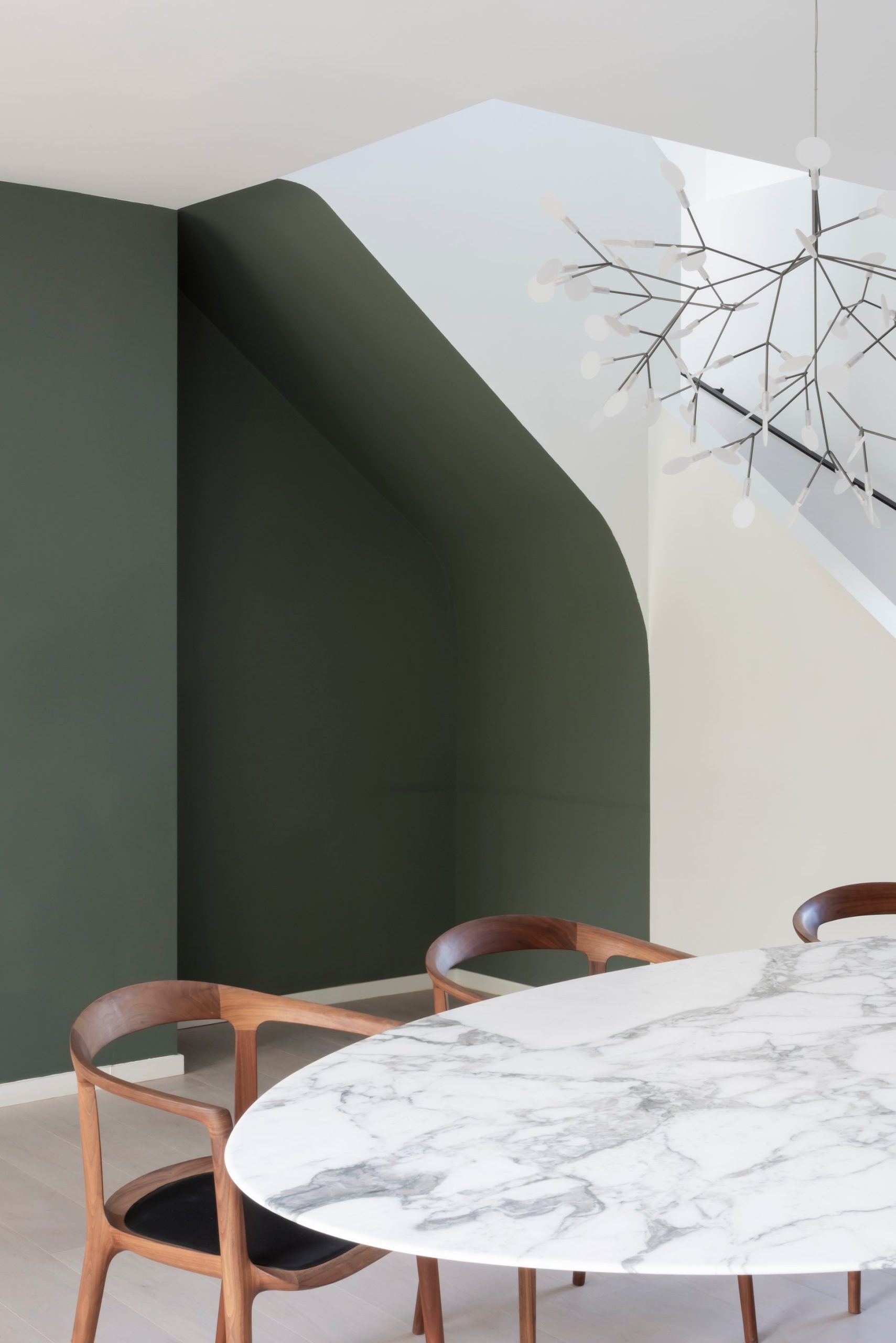 A styled dining area with an oval marble table, curved timber chairs, a deep green feature wall and a branching white pendant light beside a staircase