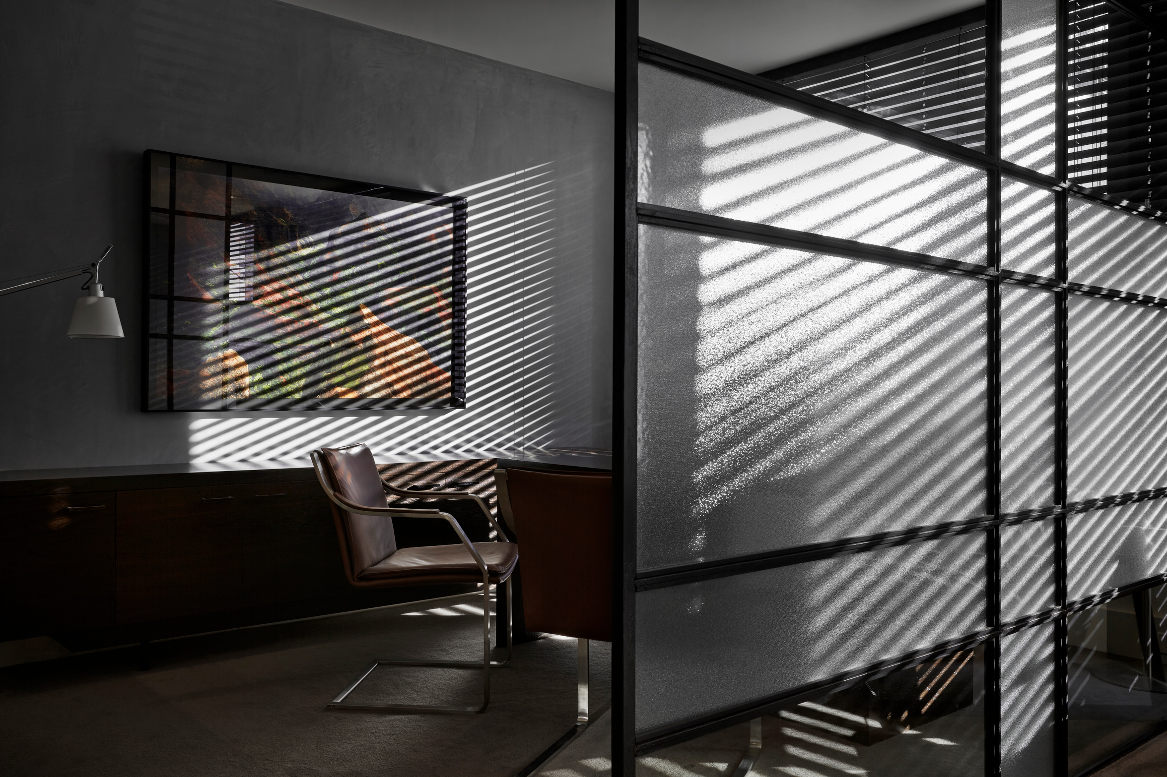 A quiet office with a leather chair, a framed artwork and a timber sideboard, striped with sunlight through venetian blinds and a steel-framed glass partition
