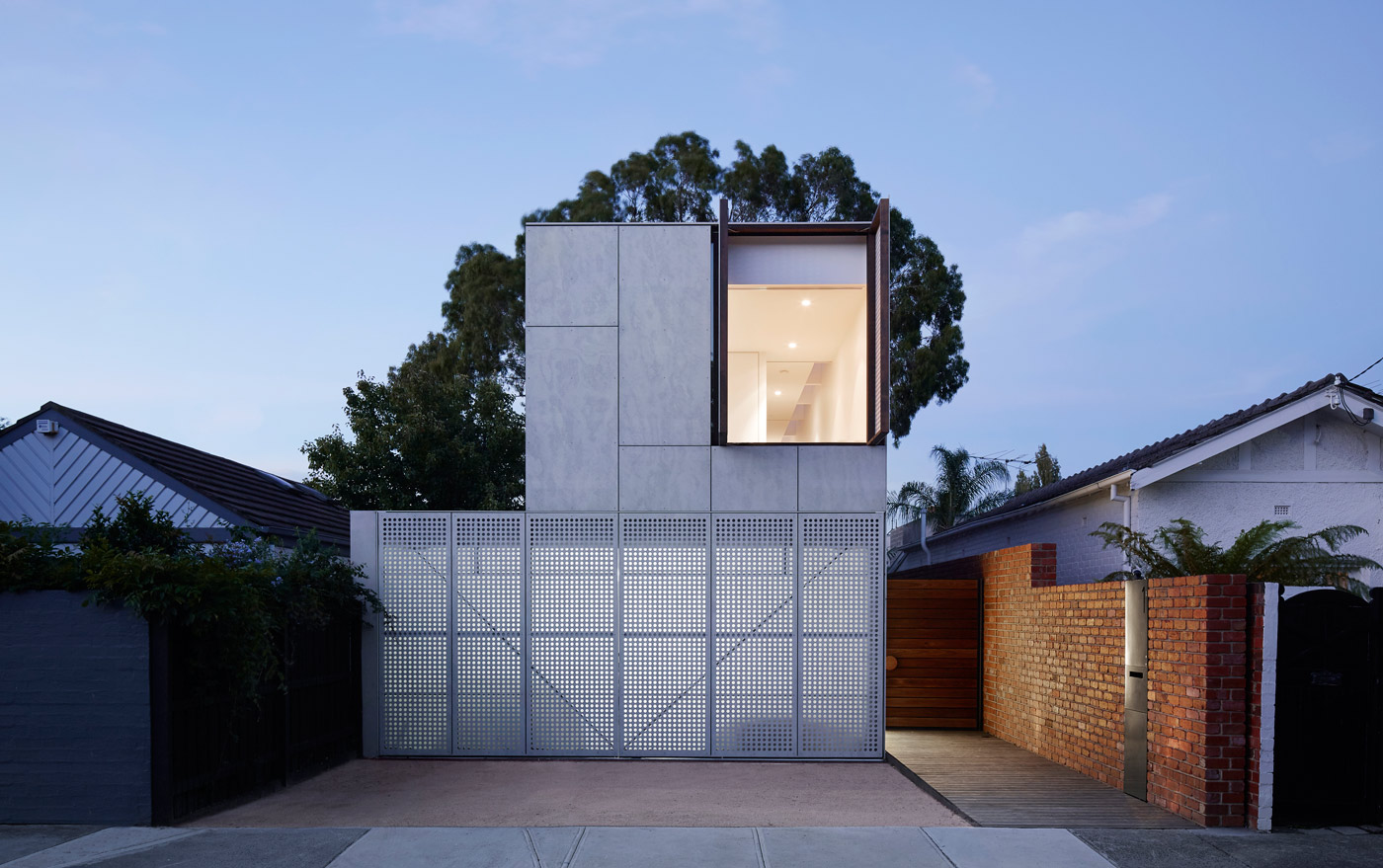 Contemporary two-storey home at dusk behind a perforated metal gate, with red brick fencing, a timber side path and a gum tree above