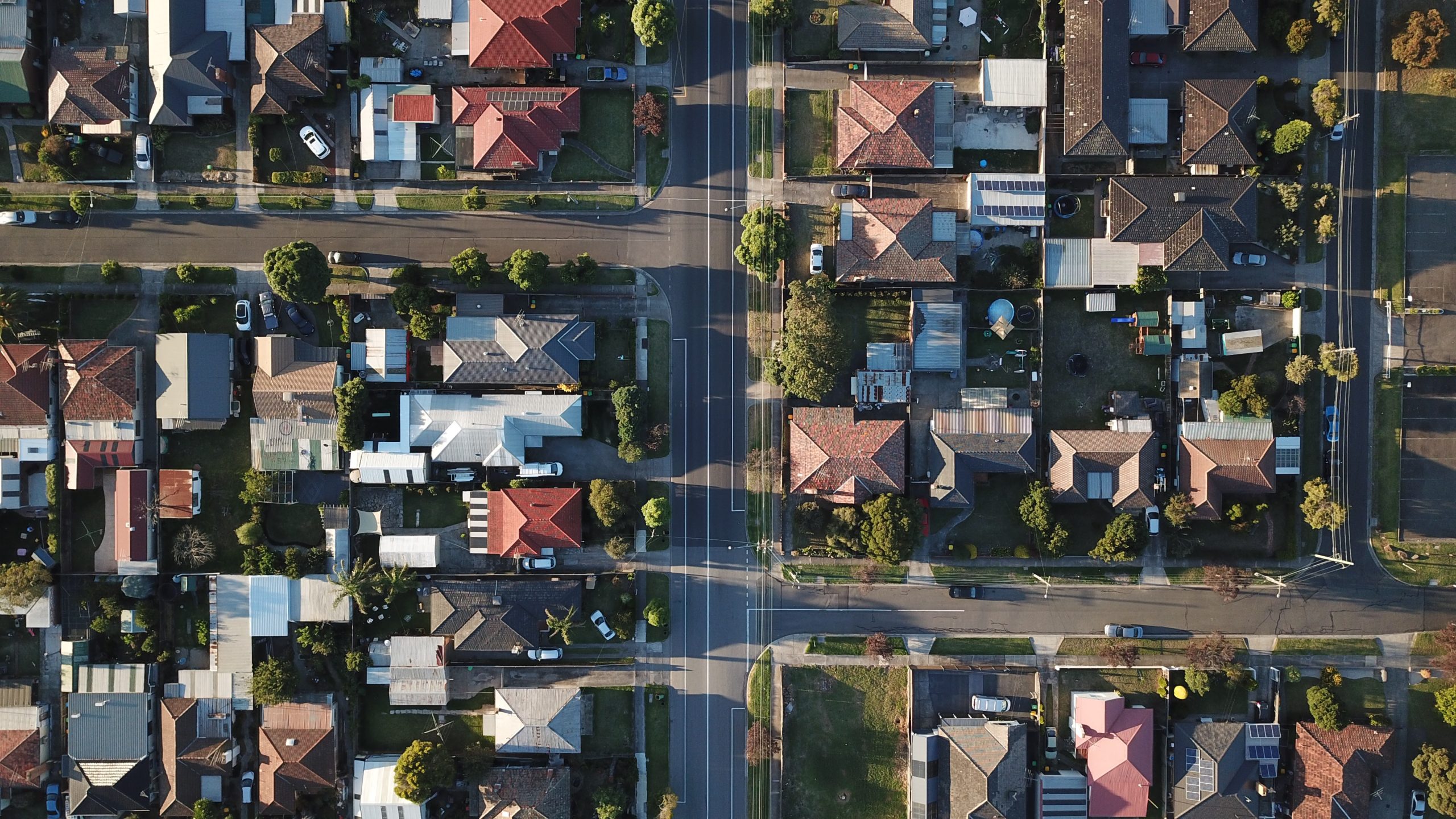 Aerial view straight down over an Australian suburban grid of tile and Colorbond roofs, backyards and tree-lined streets