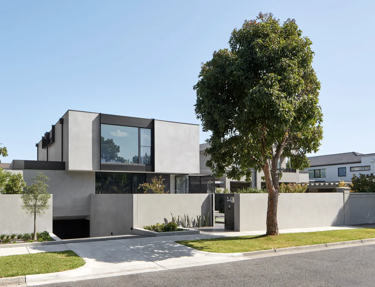 A modern two-storey rendered home behind a concrete front wall on a quiet street, with a large shade tree, a lawn verge and a clear blue sky