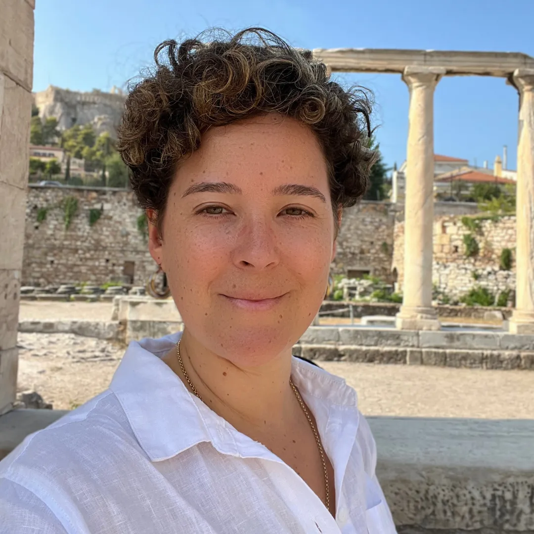 A smiling individual with short curly hair wearing a white shirt, standing in front of ancient ruins, representing a blend of modern mindfulness and historical exploration.