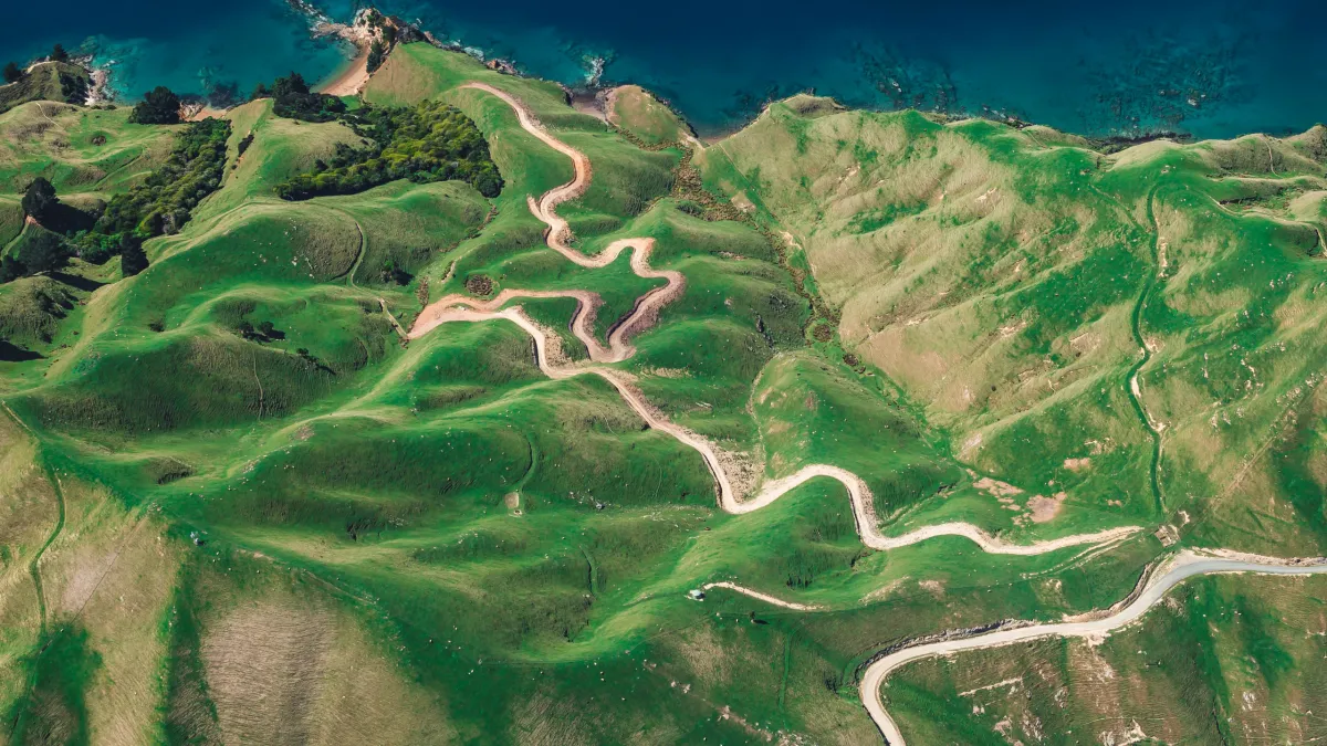 Aerial view of a lush green landscape with a winding path that weaves through rolling hills, leading to a blue shoreline at the top edge of the image.