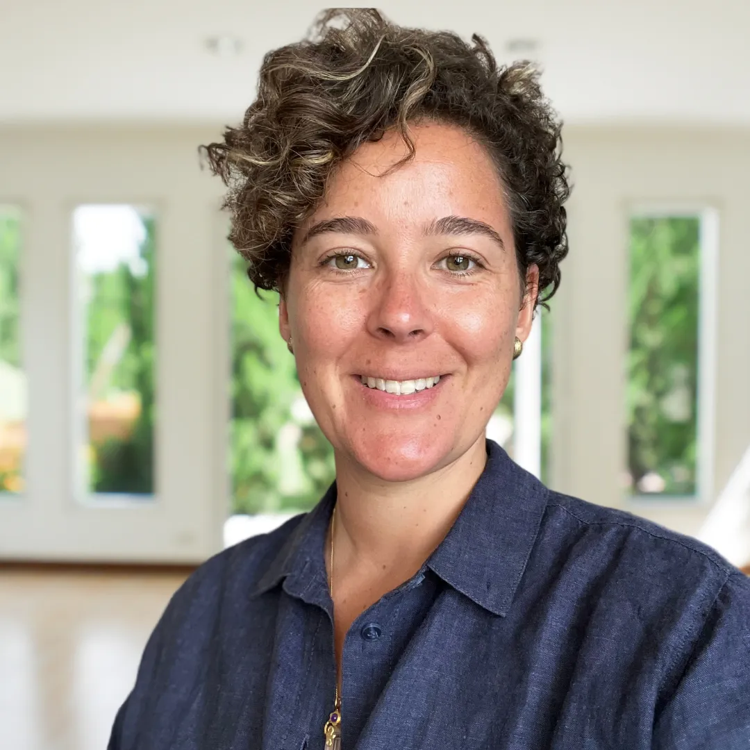 A smiling individual with short curly hair wearing a white shirt, standing in front of ancient ruins, representing a blend of modern mindfulness and historical exploration.