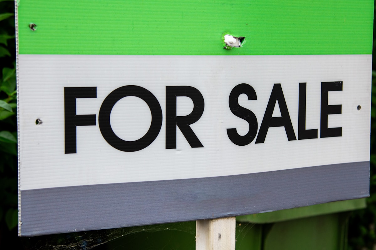 A weathered green, white, and grey FOR SALE yard sign with a small patched hole, mounted on a wooden stake in front of an East Toronto home