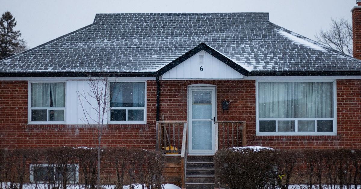 A single-storey red brick house seen from the front during a snowfall, with snow on the roof and bare hedges along the front. Wooden porch steps lead to a white storm door. The sky is overcast and no people or vehicles are visible.