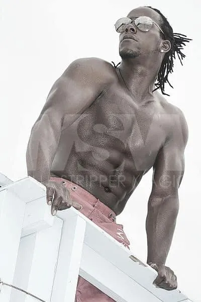 Black male stripper Kingston on a beach lifeguard tower, sporting dreads and sunglasses