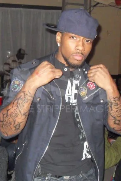 Black male stripper Pressure with a backwards navy blue cap, displaying arm tattoos