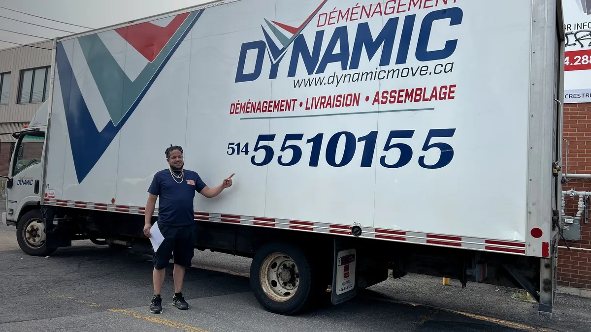 A mover in a navy uniform standing with arms crossed next to the cab of a white Dynamic moving truck.