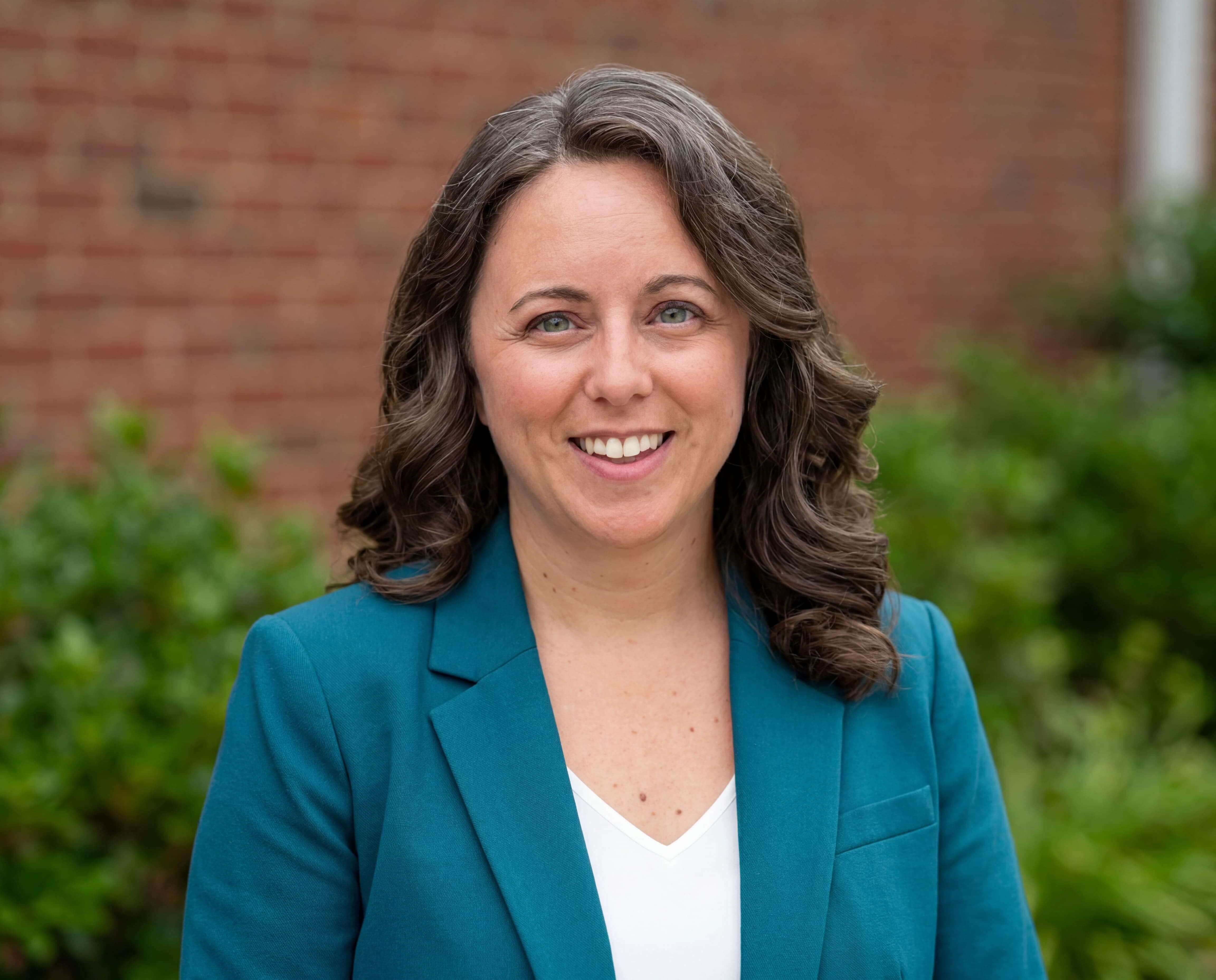 Chelsea Fournier, a legal keynote speaker, wearing a teal blazer and white blouse standing in front of a brick wall and bushes